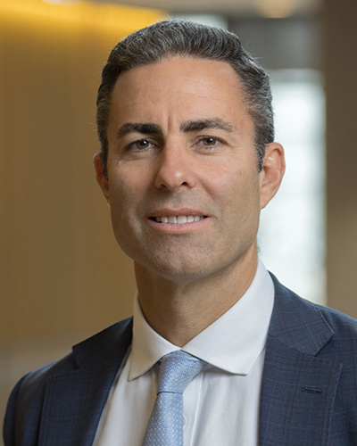 A headshot photograph of Brad smiling, looking directly at the camera, wearing a navy checkered suit, white shirt and light blue tie.