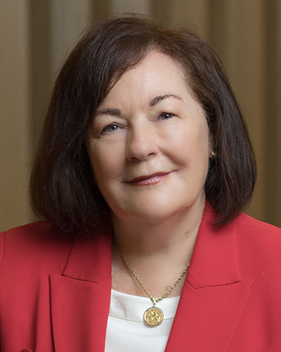 A headshot photograph of Deborah smiling, looking directly at the camera, wearing a red jacket and a white top with gold pendant necklace.