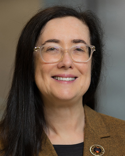 A headshot photograph of Gina smiling, looking directly at the camera, wearing clear-framed glasses, a brown tweed jacket with a round brooch on the right lapel, and a black top.