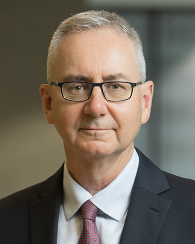 A headshot photograph of John smiling, looking directly at the camera, wearing dark-framed glasses, a black suit, white shirt and a red and grey spotted tie.