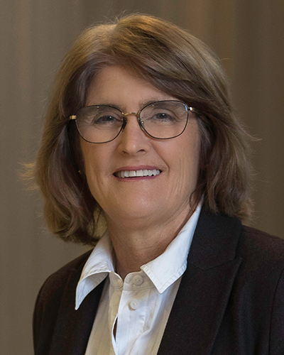 A headshot photograph of Michele smiling, looking directly at the camera, wearing dark-framed glasses, a dark jacket and a white shirt.