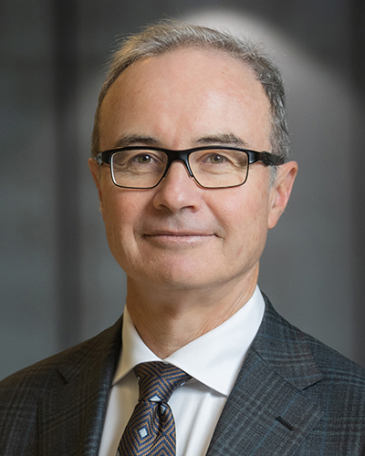 A headshot photograph of Scott smiling, looking directly at the camera, wearing dark-framed glasses, a charcoal checked suit, white shirt and a grey tie with a black and light blue diamond pattern.