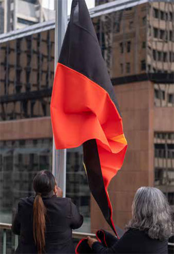 Trainee Shakeela Williams, assisted by Susan Moylan-Coombs (granddaughter of the Bank's first Governor, Dr HC Coombs), raises the Aboriginal flag at the Bank's Head Office building during National Reconciliation Week, May 2018