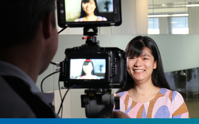 A young female economist is being filmed and we are looking at the viewpoint of the frame through the camera. She is smiling and looking directly into the lens.