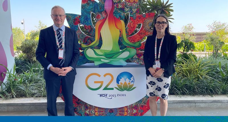 Dr Lowe and Claire are standing and smiling at the camera, standing in front of the colourful G20 signage for 2023 in India.