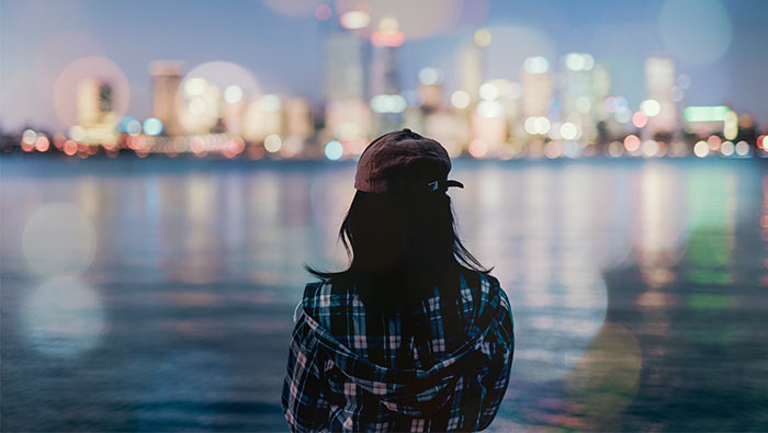 A young woman looks out across the water towards an illuminated city skyline.