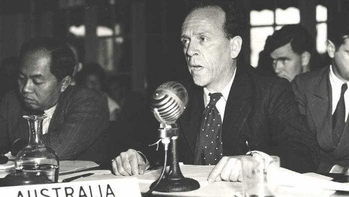 Dr HC Coombs speaking into a microphone at the Unitited Nation's Meeting for Asia and the Far East, seated at a table with a sign for Australia.