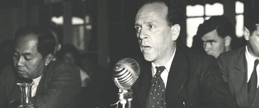 Dr HC Coombs speaking into a microphone at the Unitited Nation's Meeting for Asia and the Far East, seated at a table with a sign for Australia.