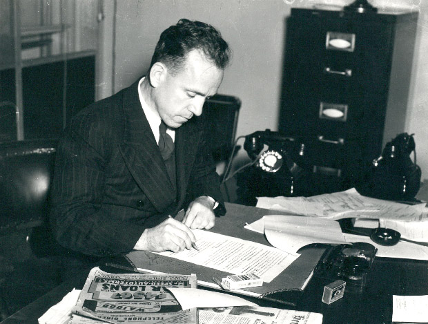 Archival photo of Dr HC Coombs as Director of Rationing, at his desk with papers and newspapers.