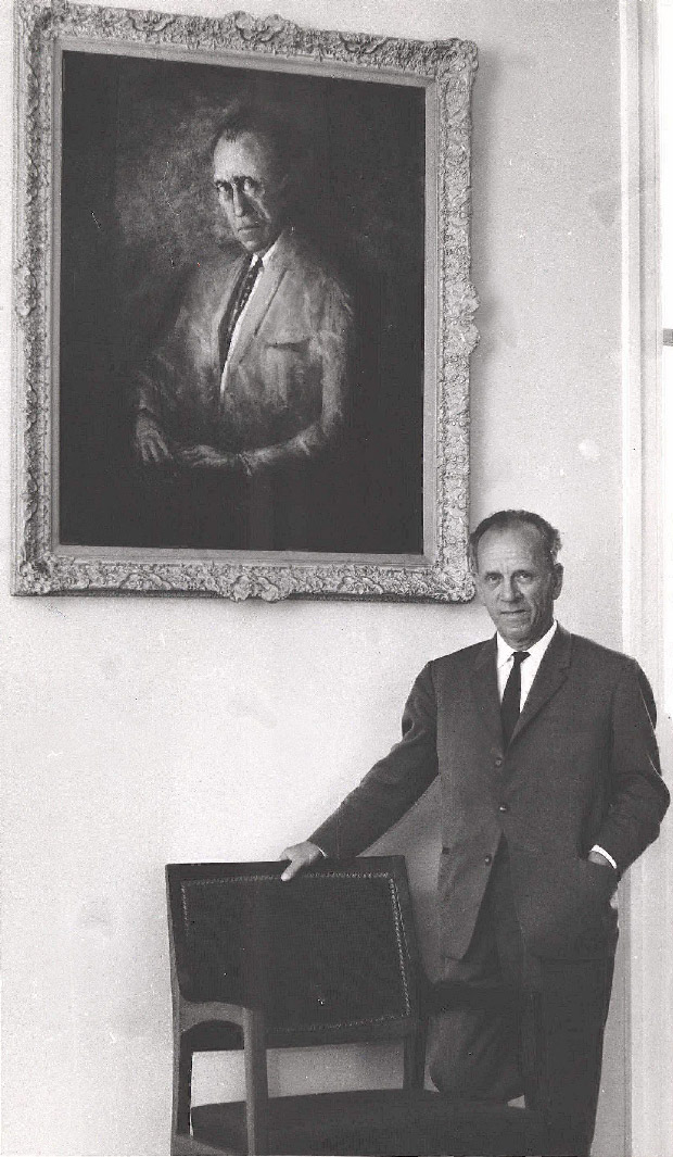 Dr HC Coombs standing in front of a portrait of himself (by Clifton Pugh) at the opening of the HC Coombs Building at ANU, with hand resting on a studded leather chair.