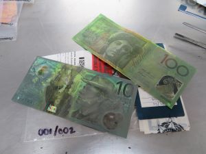One $10 and one $100 banknote with dark discolouring laid on top of a transparent plastic evidence bag containing police documents and papers on a silver table.