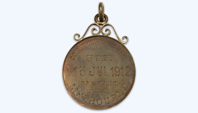 Photo of a round bronze penny with a filigree attachment at the top. The penny is inscribed with ‘Commonwealth Bank of Aust. 15 Jul 1912. Sav. Bank Dept. Melbourne’.