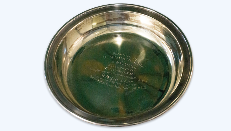 Photo of a round silver-plated dish cover. The base of the dish is inscribed with ‘Presented to G.M. Shain Esq. by J.P. Williams Salvage Ship “Claymore”. This plate was recovered from the bullion room. R.M.S. Niagara sunk in 438 feet of water at the entrance to the Hauraki Gulf N.Z.