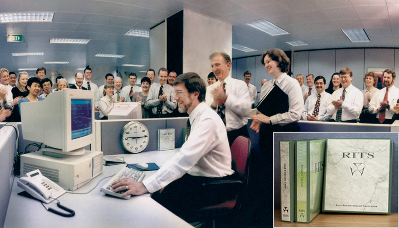 Photo showing a man with a smile on his face, in a white shirt and a tie and with glasses, sitting at an office workstation and looking at a desktop computer, with a phone on the left of the computer and a clock and a RITS manual to the right. The man is surrounded by members of staff in office attire.